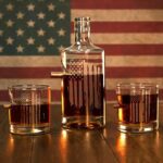 Glass decanter standing between two whiskey glasses, all embedded with a bullet and engraved with a white american flag.