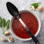 Basting spoon with steel stem and black plastic handle and spoon, lying on a bowl of tomato sauce.