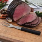 Steel carving fork with black plastic handle lying on a cutting board beside a steak cut in slices.