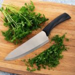 French chef knife with black plastic handle and large size blade lying on a board beside cut parsley.