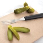 Granny fork with long tines and black plastic handle lying on cutting board beside pickles.