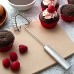 Triangular wire whisk with aluminum handle lying beside chocolate cupcakes.