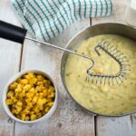 Triangular wire whisk with black plastic handle lying on a pan with macaroni and cheese.