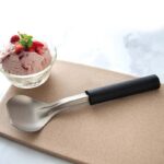 Steel ice cream scoop with black plastic handle lying beside a small bowl of strawberry ice cream.