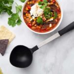 Ladle with steel stem and black plastic handle and spoon, lying beside a bowl of chilli.