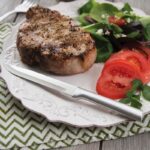 Steak knife with straight, pointy tip, steel blade and aluminum handle laying on a plate with a piece of meat and a salad.