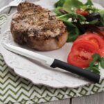 Steak knife with straight, steel blade, pointy tip and black plastic handle laying on a plate with a piece of meat and a salad.