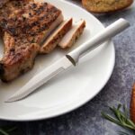 Steak knife with a serrated, steel blade, pointy tip and aluminum handle laying on a plate with a cut piece of meat.