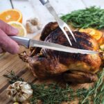 Carver knife with curved, steel blade and aluminum handle being used to cut pieces of a roasted chicken.