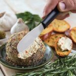 Short, broad steel knife with dull tip, slightly serrated edge and black plastic handle being used to spread soft cheese on toasts.