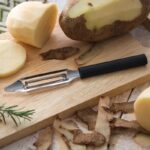 Straight peeler with swivel blade and black plastic handle lying beside peeled potatoes.