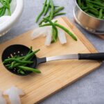 Skimmer with steel stem and black plastic handle and spoon, lying on chopping bord filled with green beans.