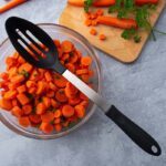 Slotted spoon with steel stem and black plastic handle and spoon, lying on bowl filled with pieces of carrot.