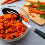 Close up of slotted spoon lying in a bowl filled with pieces of carrots and parsley.