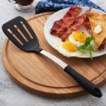 Slotted turner with steel stem and black plastic handle and face, lying beside bacon and egg breakfast.