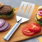 Broad spatula with aluminum handle and three slots in the face, lying beside prepared burgers.