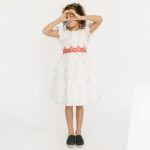 Young girl wearing white dress decorated with colored dots, embroidery along the waist and flutter sleeves.