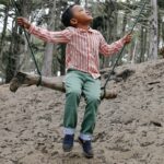 Kid sittingon swing wearing green trousers and pink- and white striped shirt.