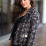 Woman wearing tweed blazer in black and pastel color palette, with rosette buttons and fringed trims.