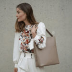 Woman carrying large dark beige square leather bag with strap.